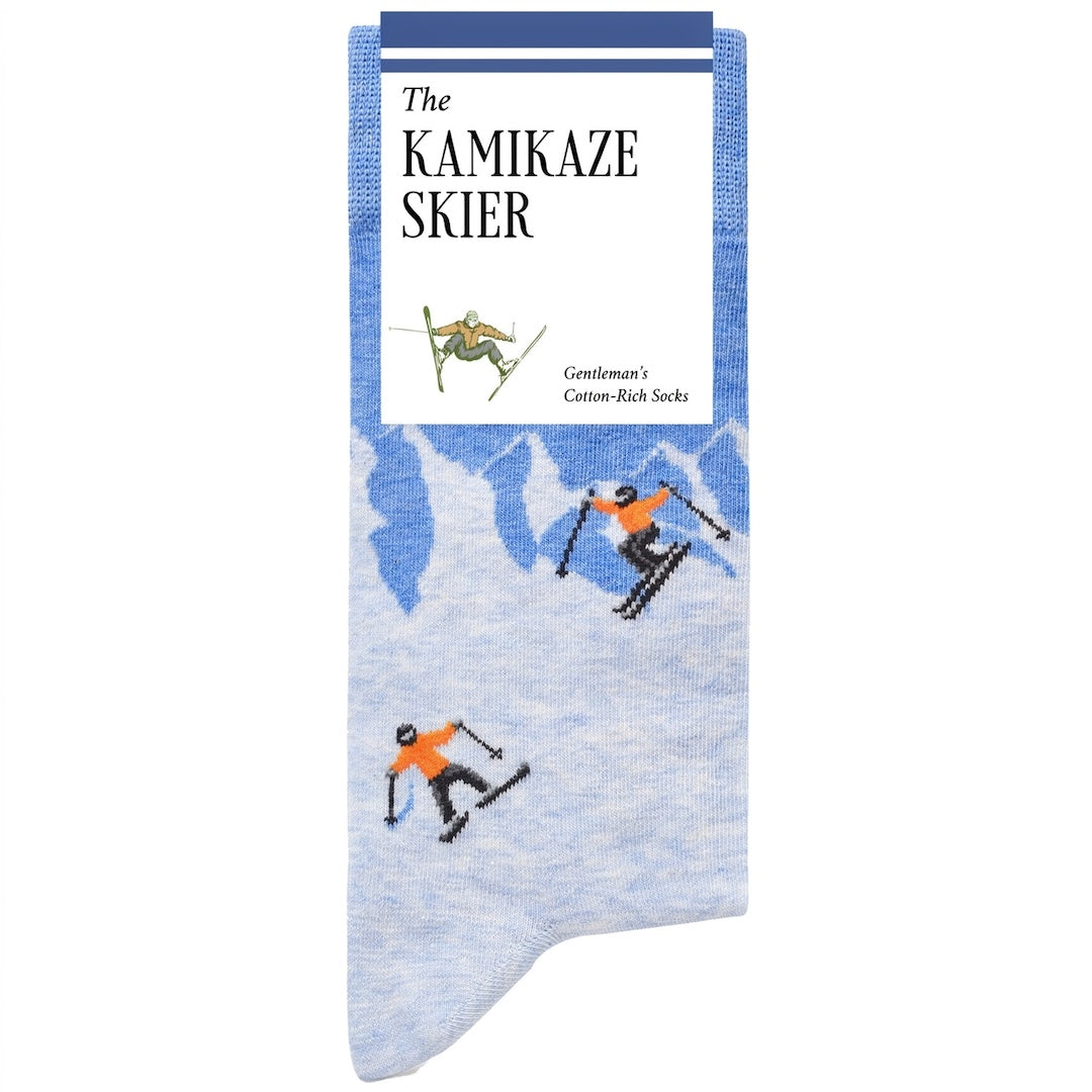A pair of knit socks with a light blue and white mountain pattern and skiers printed on them, featuring a white paper header card that reads 'The KAMIKAZE SKIER'.