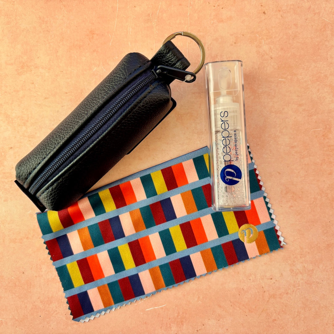 A navy blue faux-leather zipper pouch with a key ring, a clear bottle of Peepers eye glasses cleaner, and a microfiber cleaning cloth with a colorful striped pattern, arranged on a peach-colored textured background.
