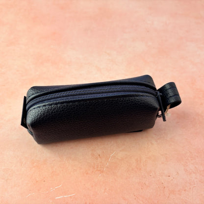 A small navy blue pebbled leather zip pouch with a top zipper and a small attached loop, photographed on a warm peach-pink textured surface.