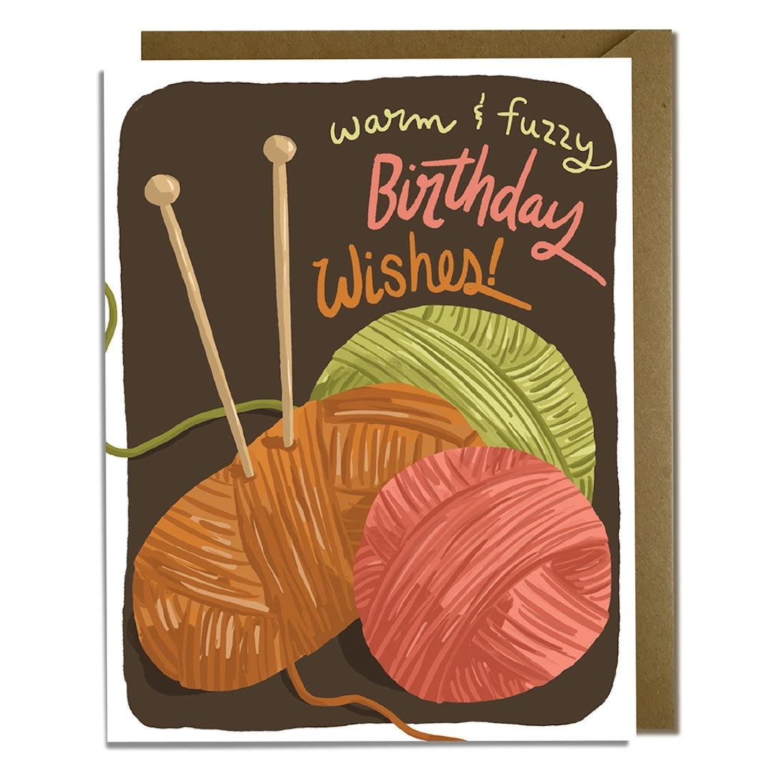 Greeting card with yarn balls and knitting needles on a brown background with text that says "warm and fuzzy birthday wishes"