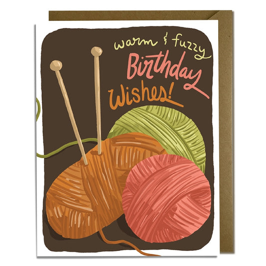 Greeting card with yarn balls and knitting needles on a brown background with text that says "warm and fuzzy birthday wishes"