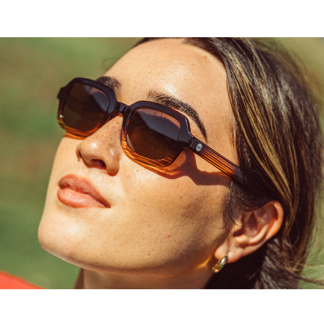 A close-up lifestyle shot of a woman wearing dark brown square sunglasses with amber-tinted lenses and tortoiseshell-patterned temples, set against a blurred green background.