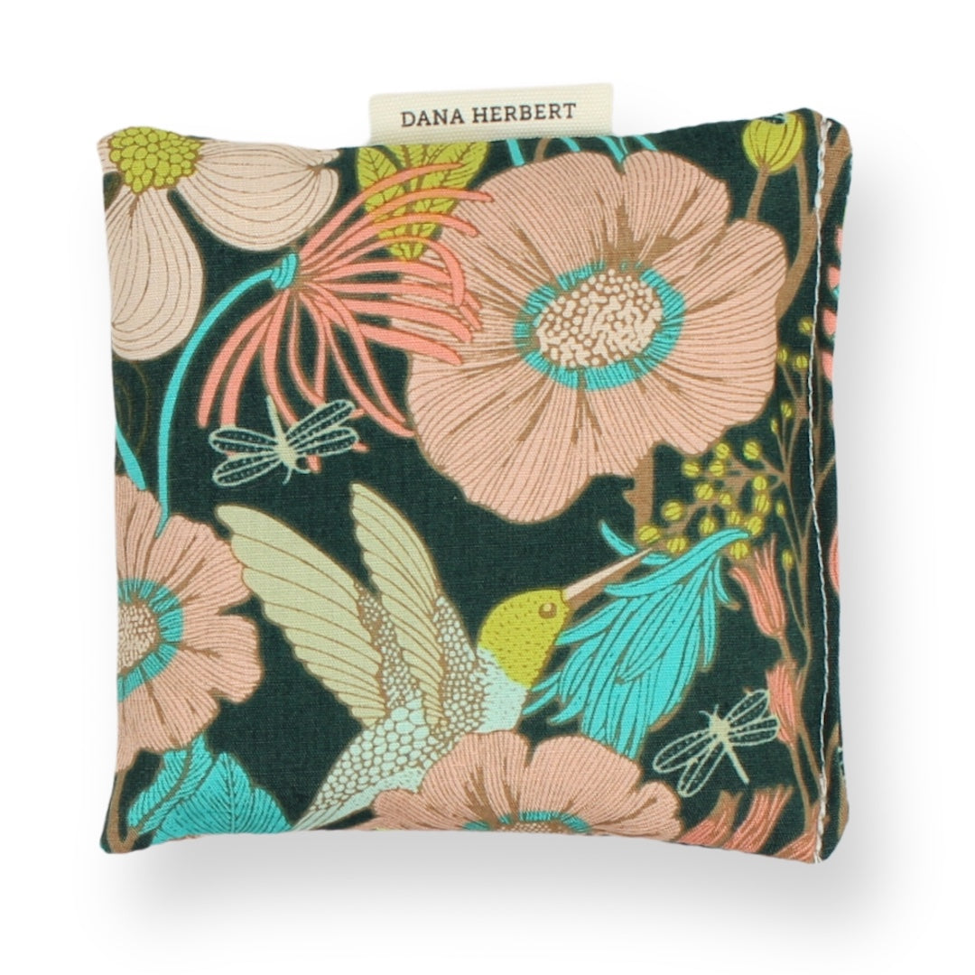 A square lavender sachet featuring a vibrant floral and bird pattern on a dark green background, with a small fabric label reading 'DANA HERBERT' at the top.