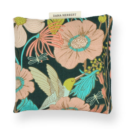 A square lavender sachet featuring a vibrant floral and bird pattern on a dark green background, with a small fabric label reading 'DANA HERBERT' at the top.