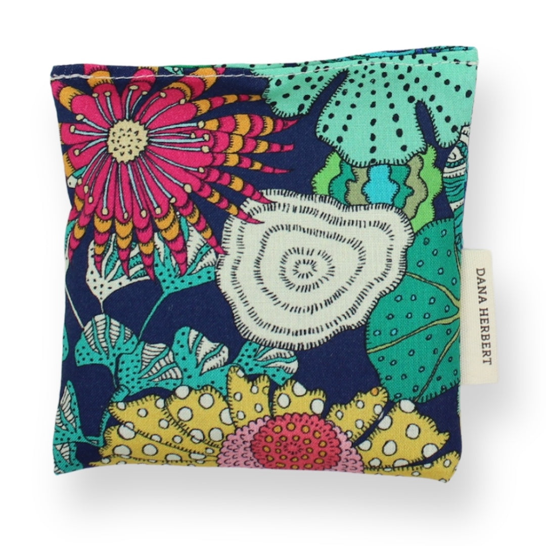 A square lavender sachet featuring a vibrant, multicolored floral and botanical pattern on a dark navy background. The pattern includes bright pink, yellow, teal, green, and white elements.
