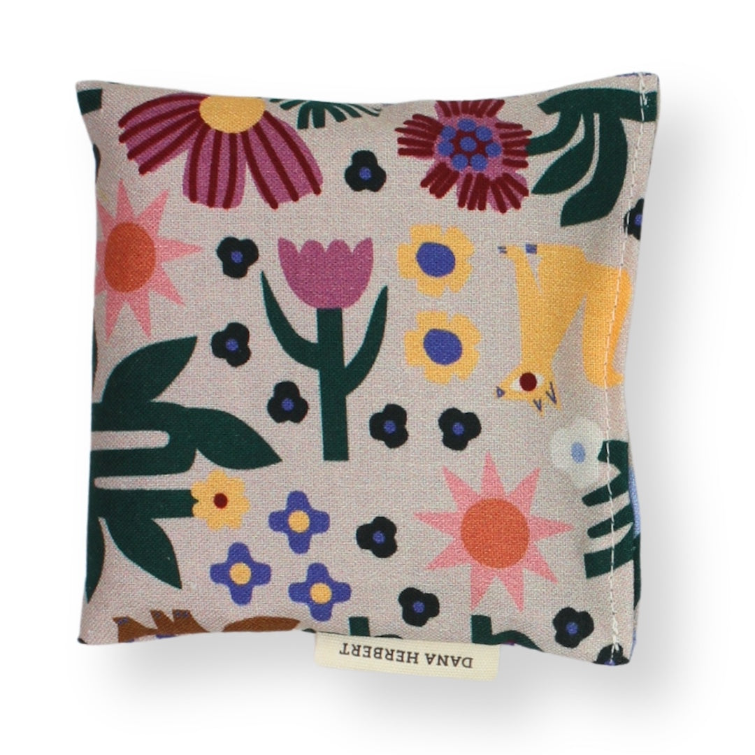 A square lavender sachet featuring a colorful, whimsical floral and animal pattern on a light beige linen-like fabric. The pattern includes stylized flowers, leaves, and a yellow bird.