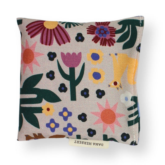 A square lavender sachet featuring a colorful, whimsical floral and animal pattern on a light beige linen-like fabric. The pattern includes stylized flowers, leaves, and a yellow bird.