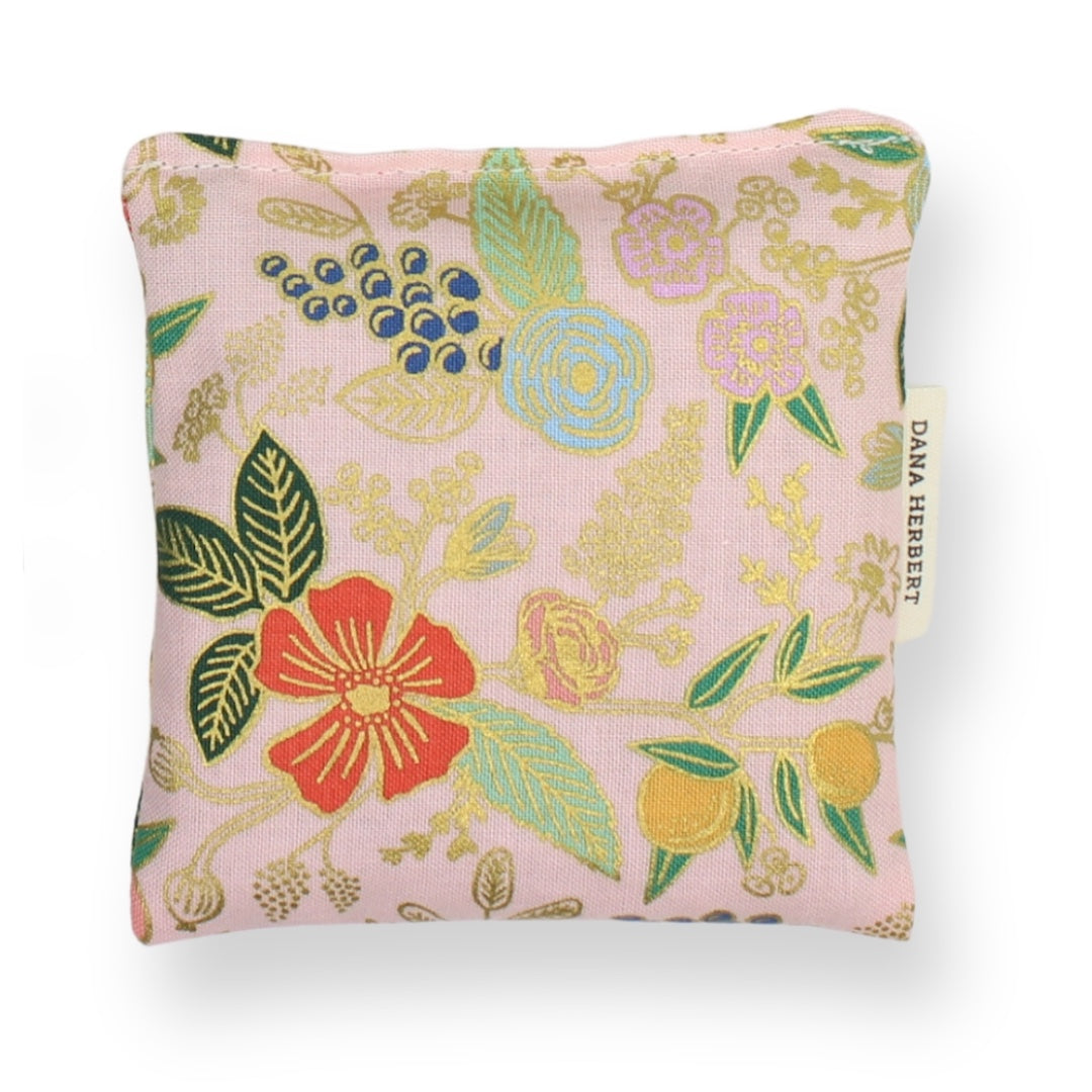 A square lavender sachet, featuring a light pink linen-like fabric printed with a botanical pattern in gold, green, blue, and red-orange, with a small white brand tag on the right side.