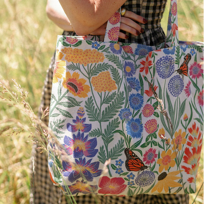 A white tote bag with a colorful wildflower and butterfly print, held by a person in a grassy outdoor setting.
