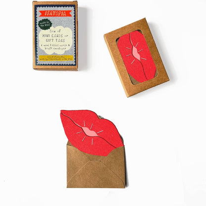 Red lip shaped card  inside a brown envelope, next to its packaging box on a white background.