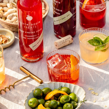 Bottles of Little Apple Treats cider vinegar shrub with cocktails and garnishes on a table.
