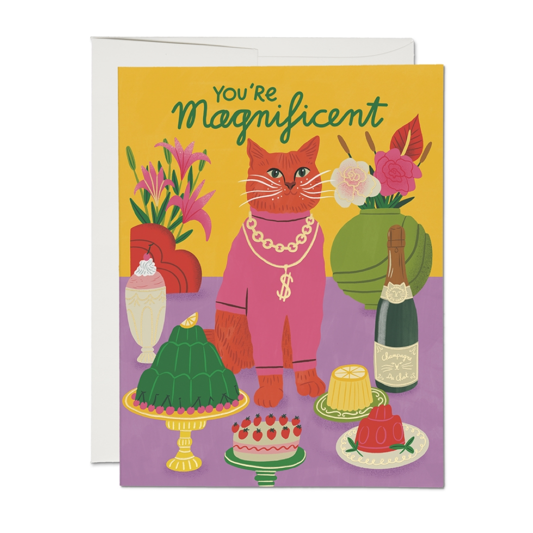 A greeting card with a white envelope behind it, featuring a colorful illustration of an orange cat wearing pink clothing and gold jewelry, surrounded by desserts, flowers, and a champagne bottle. The card reads 'You're Magnificent' in green text on a yellow background.