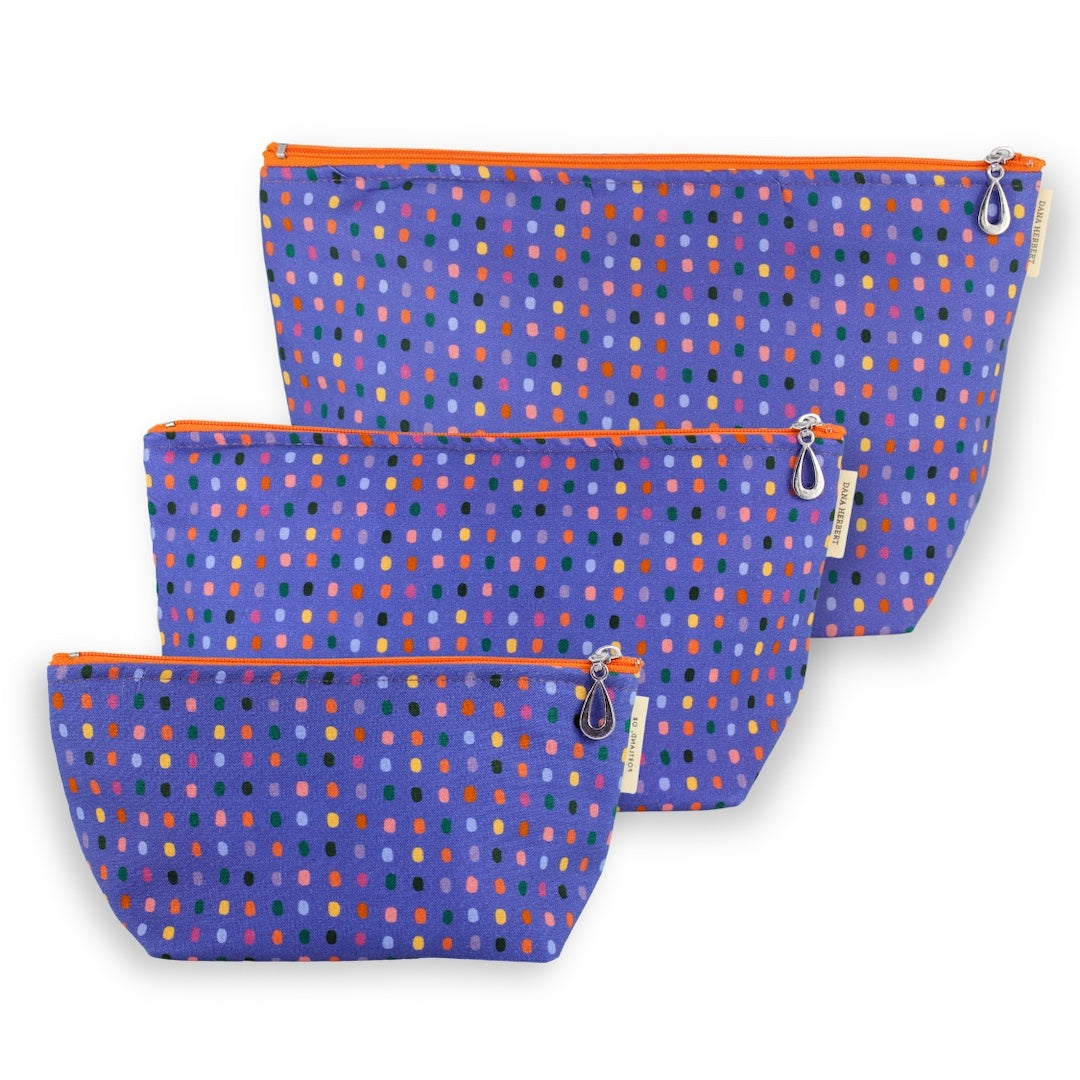 Three fabric zip pouches in a nested set, featuring a purple background with a multicolor dotted pattern and bright orange zippers.