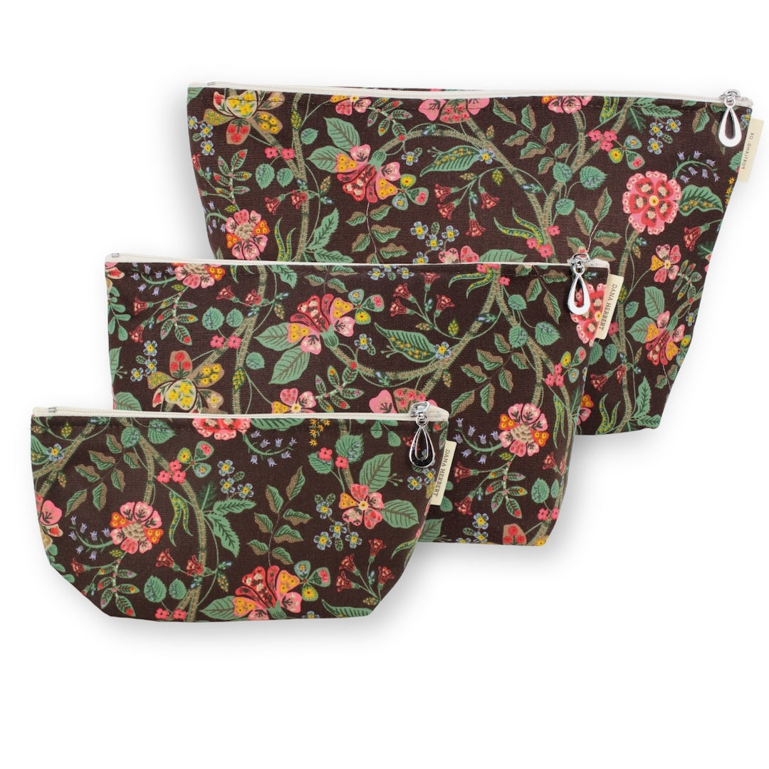 Three floral zippered pouches in different sizes, arranged diagonally on a white background. The pouches feature a dark brown base with a vibrant multicolor botanical pattern of pink flowers, green leaves, and yellow accents.