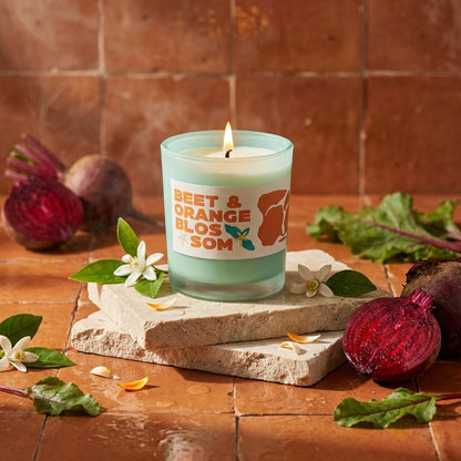Candle in a glass vessel with a label that says 'beet and orange blossom' on a stone block surrounded by beets and flowers on a wooden surface.