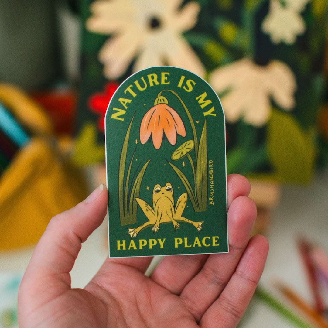 Hand holding a sticker with a frog sitting in a garden scape with text that reads nature is my happy place