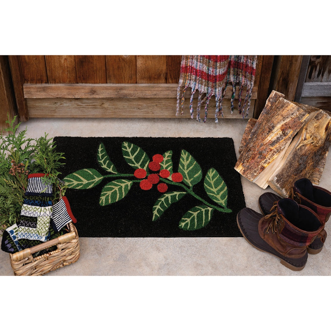 A rectangular black doormat with a green holly leaf and red berry design, styled on a concrete porch with a wooden fence, folded wood, boots, and a basket of knit items.