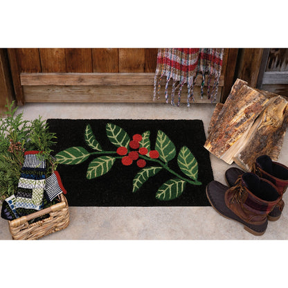A rectangular black doormat with a green holly leaf and red berry design, styled on a concrete porch with a wooden fence, folded wood, boots, and a basket of knit items.
