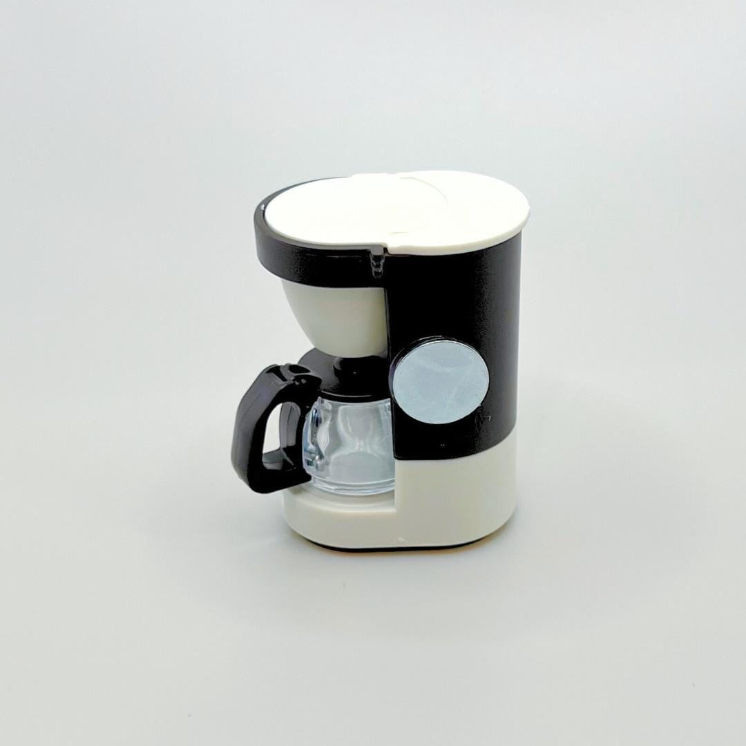 A small coffee maker with a black and off-white body, featuring a clear glass carafe, black handle, and a circular magnet, set against a plain light gray background.