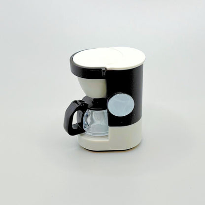 A small coffee maker with a black and off-white body, featuring a clear glass carafe, black handle, and a circular magnet, set against a plain light gray background.