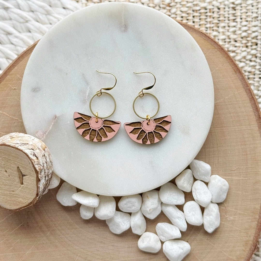 Pair of earrings with a brass circle and half circle pieces of wood that have been painted pink and have a flower pattern carved out on a marble plate with pebbles and wood slice in the background
