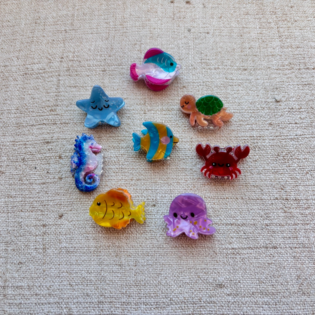 A set of eight colorful, cartoon-style sea creature hair clips (starfish, turtle, seahorse, fish, crab, octopus) arranged on a textured beige linen fabric background.
