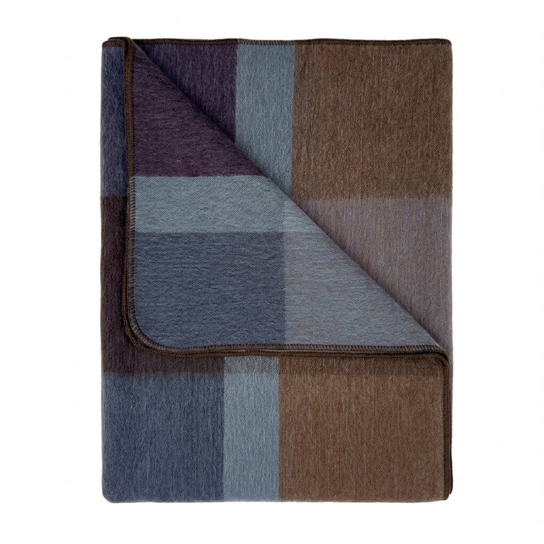 A folded plaid blanket with a textured woven surface, featuring blocks of brown, slate blue, lavender purple, and gray, shown on a plain white background.