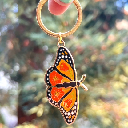 A butterfly-shaped keychain charm with orange, yellow, black, and white enamel details, attached to a gold-tone split ring and held by a hand against a blurred outdoor background.