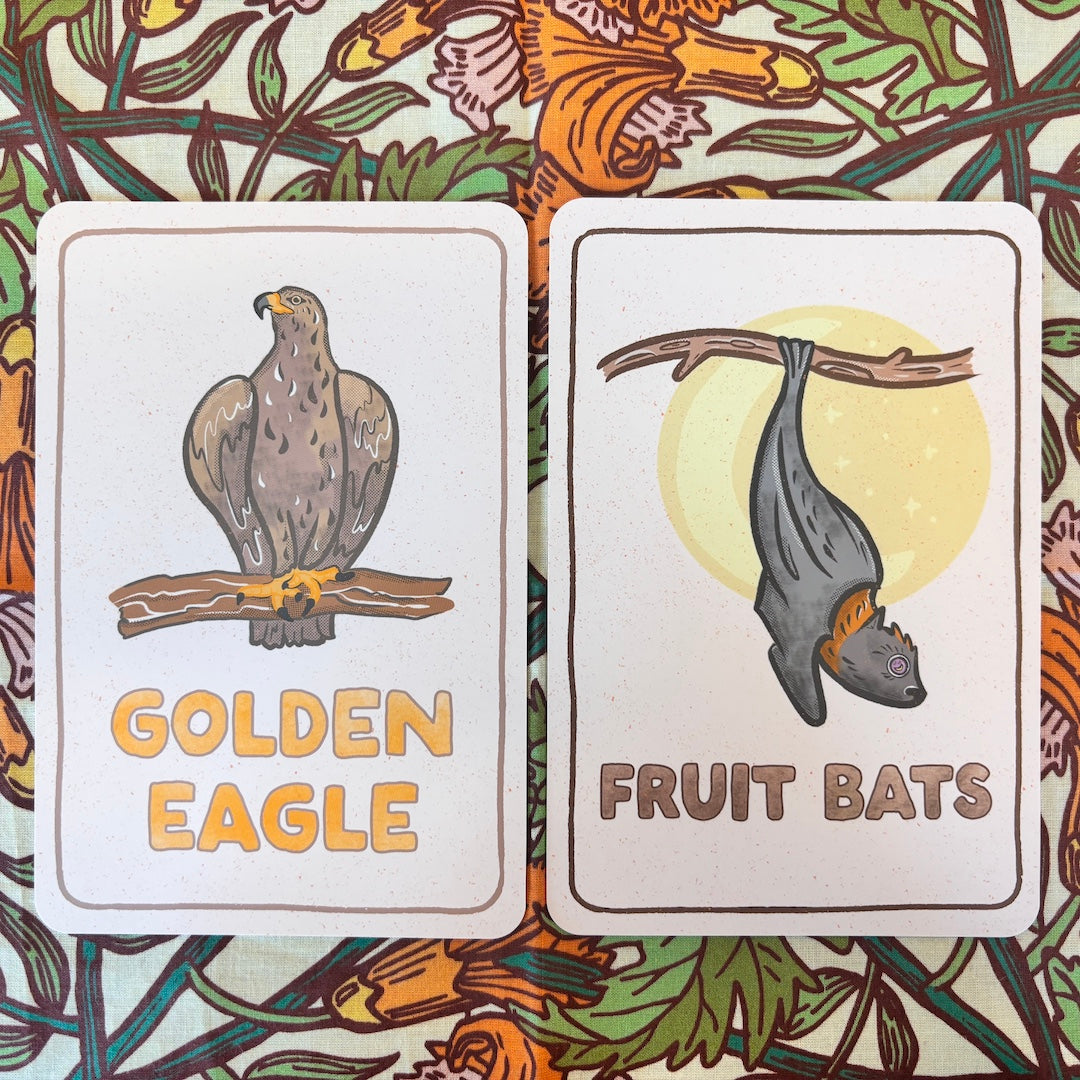 Two illustrated cards laid side by side on a floral patterned surface. The left card shows a golden eagle perched on a branch with the text 'GOLDEN EAGLE.