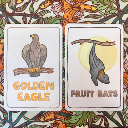 Two illustrated cards laid side by side on a floral patterned surface. The left card shows a golden eagle perched on a branch with the text 'GOLDEN EAGLE.