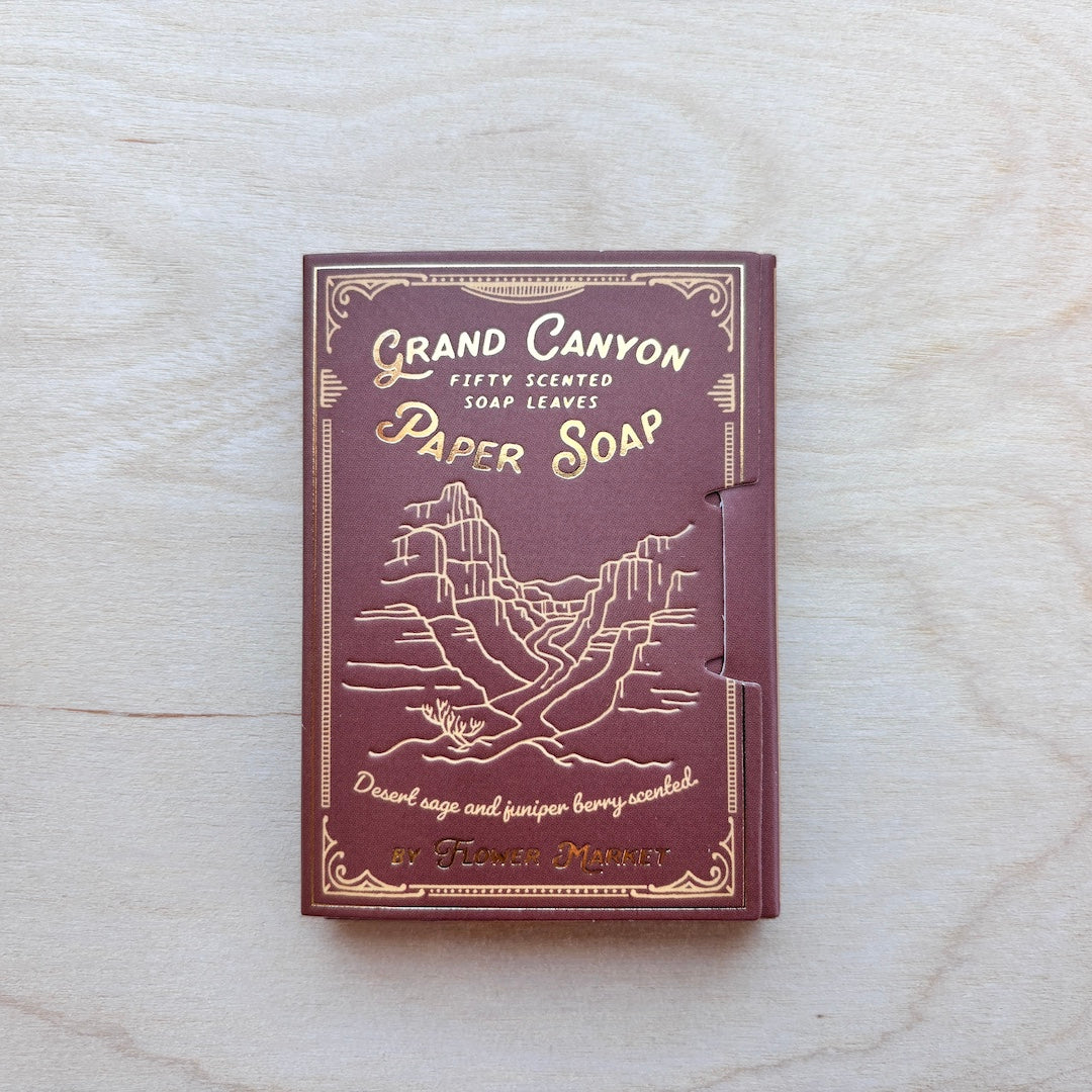 A small rectangular box of Grand Canyon Fifty Scented Soap Leaves paper soap, featuring a maroon cover with gold-embossed desert canyon artwork and lettering, placed on a light wood surface.