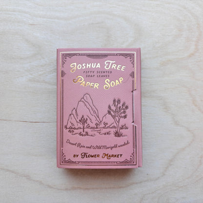 A pink box of Joshua Tree Paper Soap by Flower Market, featuring gold foil lettering and desert landscape illustrations, placed on a light wood surface.