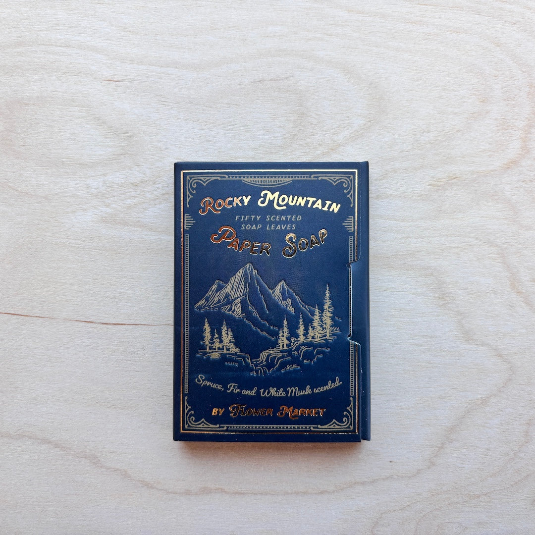 A vintage-style soap bar package labeled 'Rocky Mountain Paper Soap' by Helen Harvey, shown standing upright on a light wood surface. The package is dark blue with gold-toned illustrated mountain scenery and lettering.