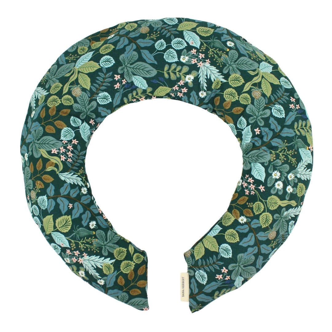 A padded neck pillow with a detailed botanical pattern featuring green, teal, and brown leaves with small white and pink flowers on a dark teal background. A small fabric tag is visible at the bottom right.