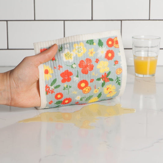 Hand holding a floral-patterned swedish dishcloth over a spill on a countertop with a glass of orange juice.