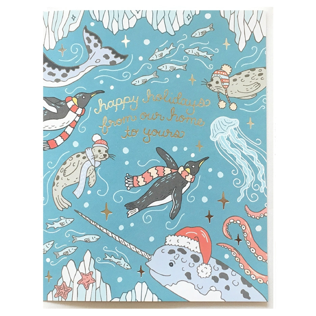 A square holiday greeting card featuring an underwater winter scene with penguins, seals, a narwhal, octopus, jellyfish, fish, and stars on a light blue background. Gold script reads 'happy holidays from our home to yours.