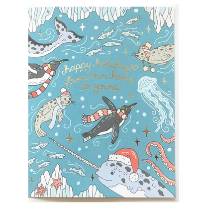 A square holiday greeting card featuring an underwater winter scene with penguins, seals, a narwhal, octopus, jellyfish, fish, and stars on a light blue background. Gold script reads 'happy holidays from our home to yours.