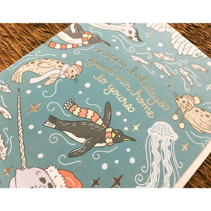 A square holiday card featuring a light blue background with whimsical illustrations of penguins, seals, a jellyfish, a unicorn, and gold script reading 'Happy Holidays from our home to yours'. The card is photographed at an angle on a dark wood surface.