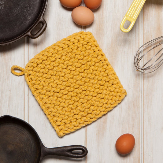 Yellow knitted pot holder on a wooden surface with eggs and skillets.
