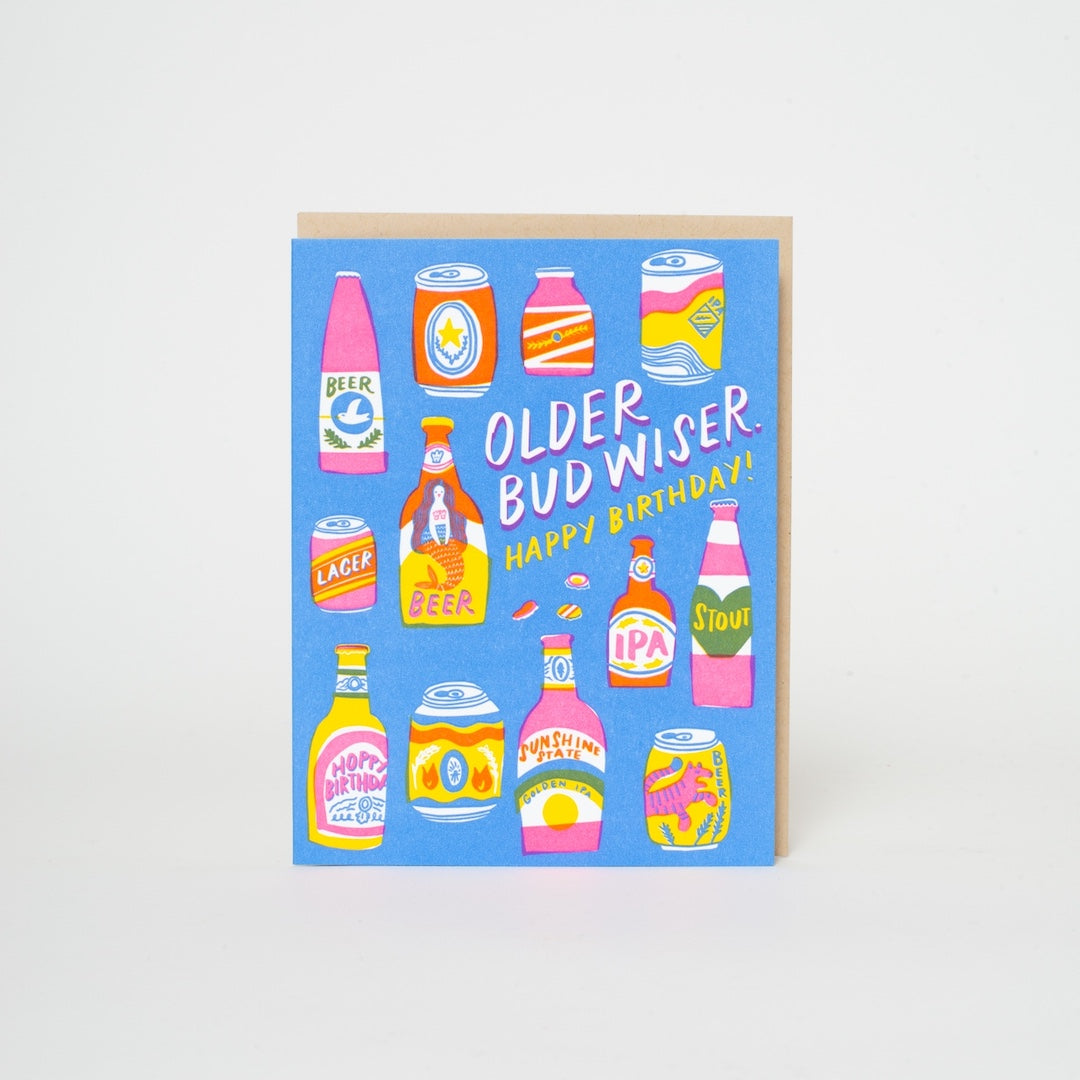 A square birthday card with a light blue background featuring colorful illustrated beer bottles and cans, with the text 'OLDER BUD WISER. HAPPY BIRTHDAY!' in white and yellow.