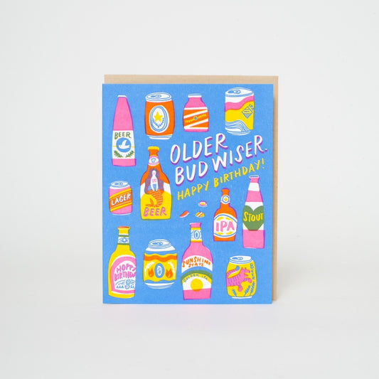 A square birthday card with a light blue background featuring colorful illustrated beer bottles and cans, with the text 'OLDER BUD WISER. HAPPY BIRTHDAY!' in white and yellow.