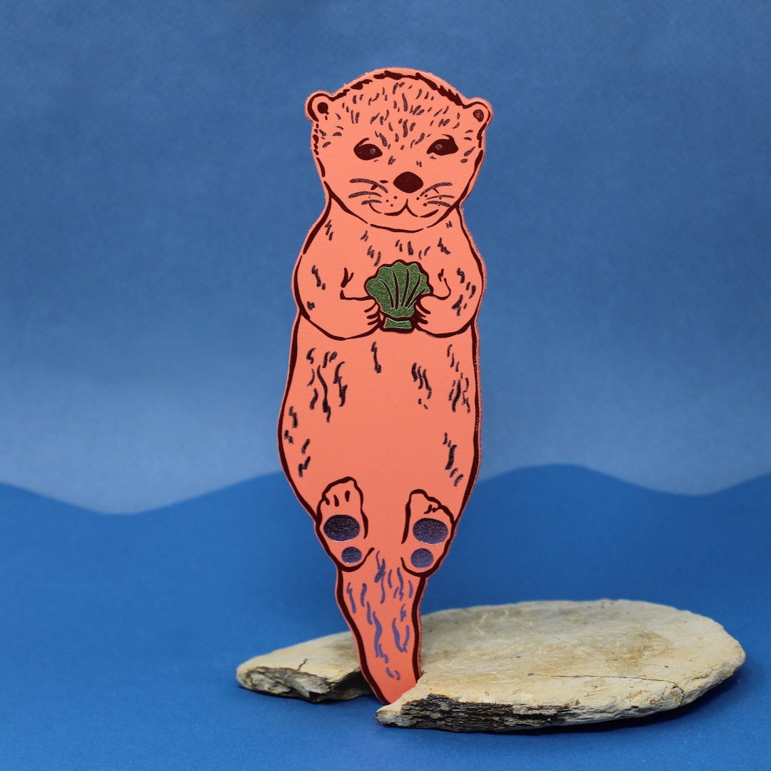apricot colored bookmark that is shaped like an otter holding a shell on a blue background