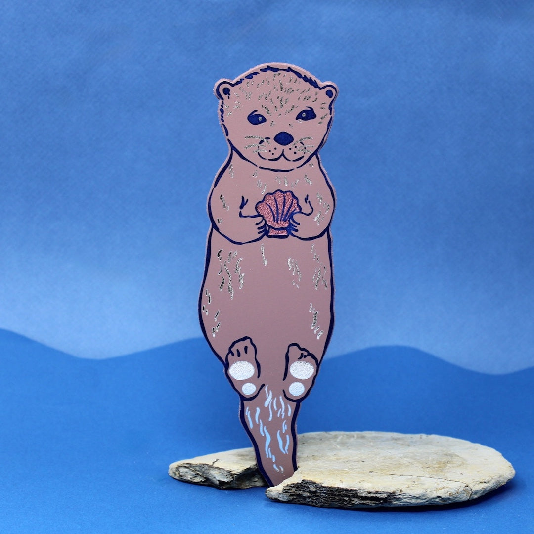 dusty pink bookmark that is shaped like an otter holding a shell on a blue background