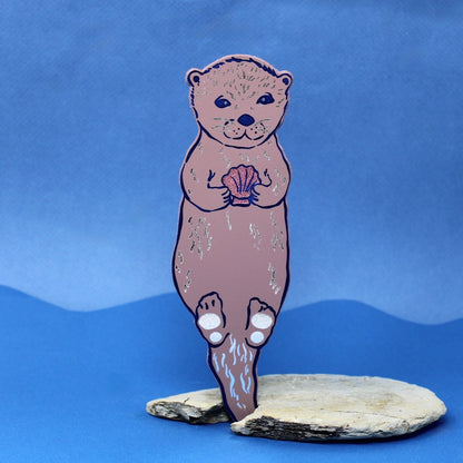 dusty pink bookmark that is shaped like an otter holding a shell on a blue background