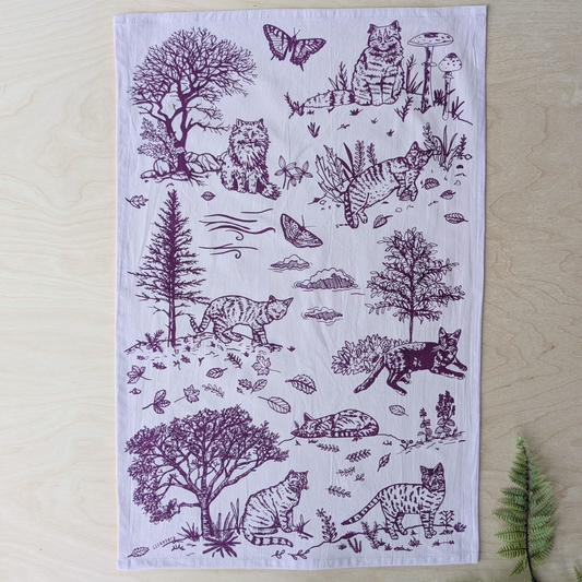 A rectangular white fabric tea towel with a dark purple printed design of various cats, trees, and butterflies, laid flat on a light wood background.