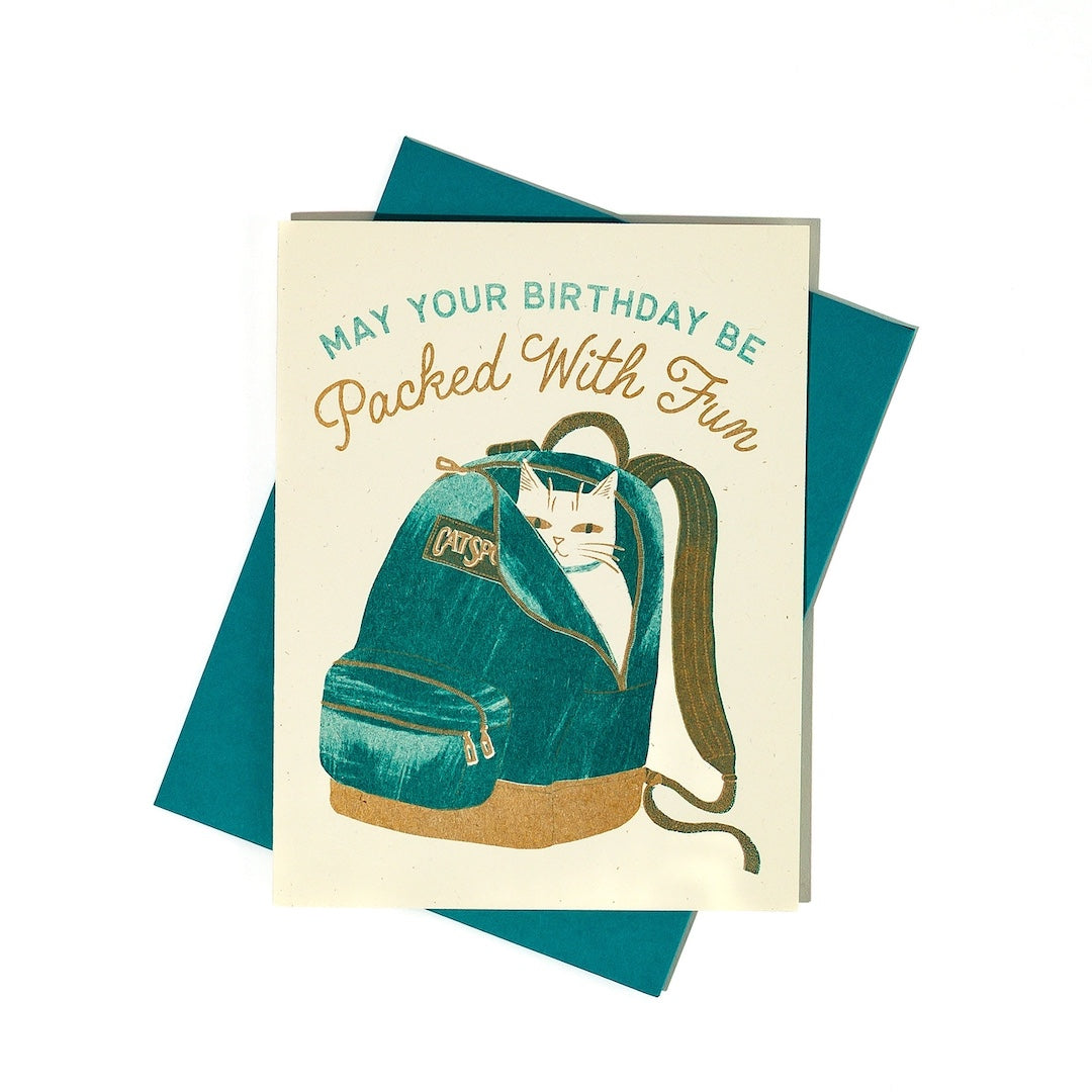 Greeting card with a white cat in a teal backpack illustration with "may your birthday be packed with fun" written on it on a white background