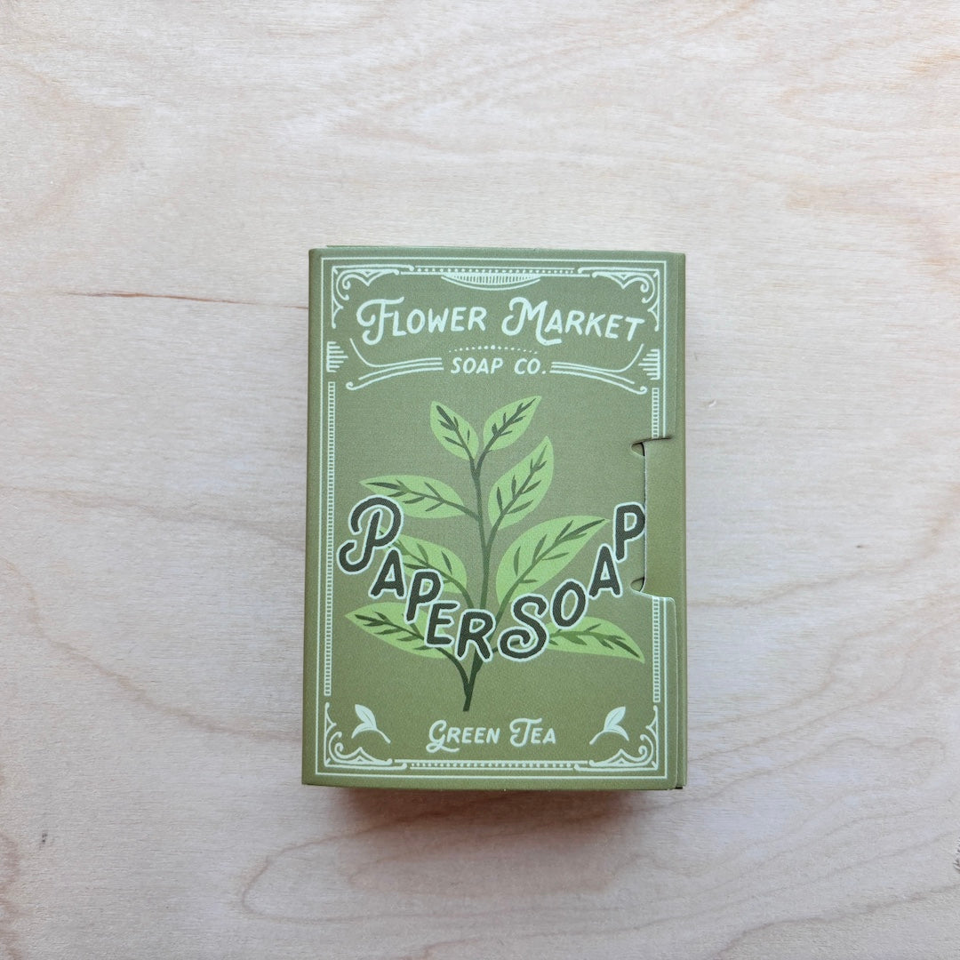 A rectangular box of Flower Market Soap Co. Paper Soap in Green Tea scent, featuring a green and white illustrated design, placed on a light wood grain surface.