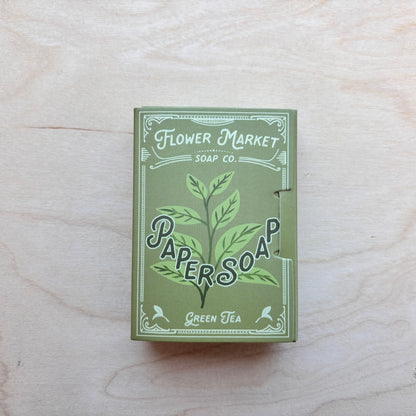 A rectangular box of Flower Market Soap Co. Paper Soap in Green Tea scent, featuring a green and white illustrated design, placed on a light wood grain surface.