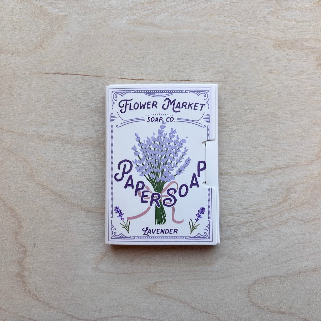 A boxed bar of Lavender Papers Soap by Flower Market Soap Co., featuring a white box with a purple lavender illustration, placed on a light wood grain surface.