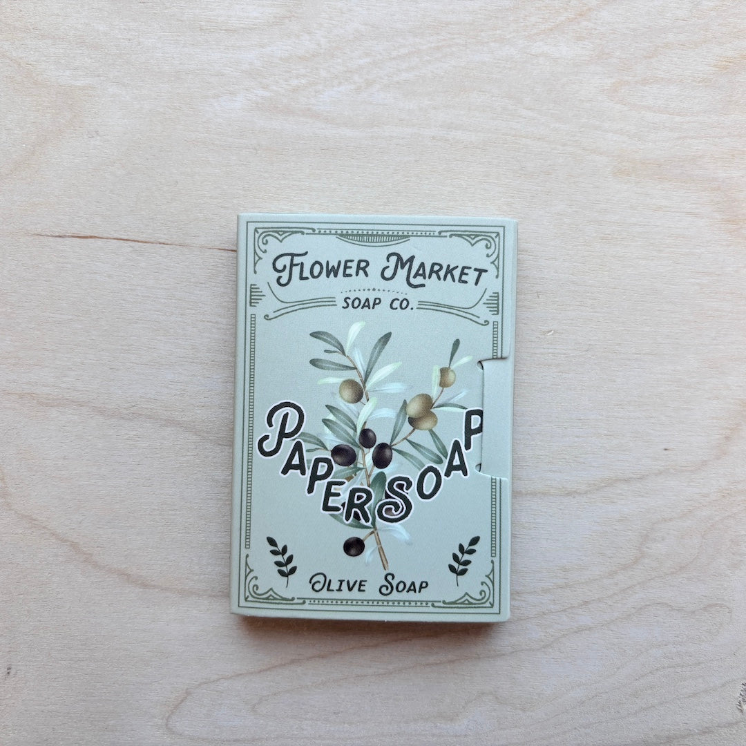 A square box of Paper Soap Olive Soap by Flower Market Soap Co. lies on a light wood grain surface.